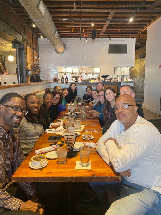 Fourteen people sit around a long dinner table with food on it, smiling.