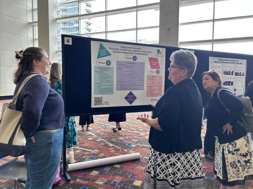 People gather around a research poster during the ALA Annual Conference, listening as a presenter discusses findings about the role of law libraries in serving system-impacted communities. Large windows and additional poster presentations are visible in the background.