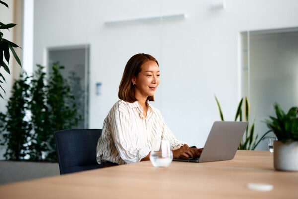 Woman working on a laptop at a conference table in a bright office with plants in the background.