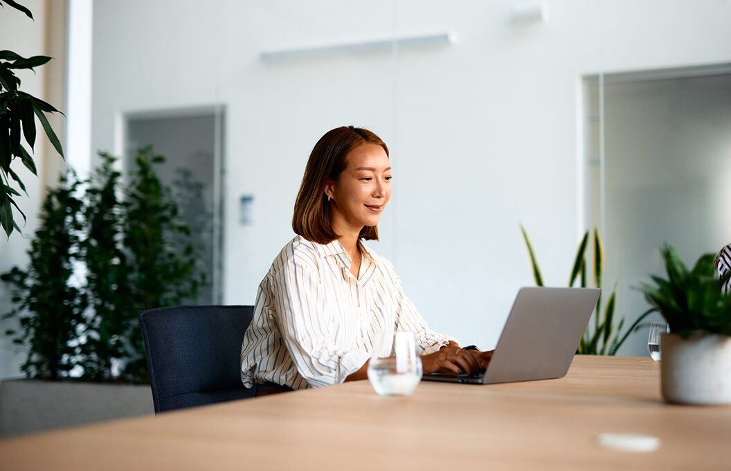 Woman working on a laptop at a conference table in a bright office with plants in the background.