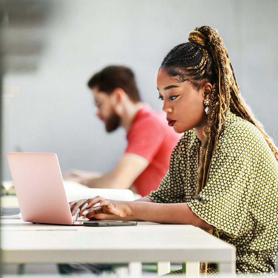 Woman with braided hair typing on a laptop in the foreground, with a blurred colleague seated behind her.