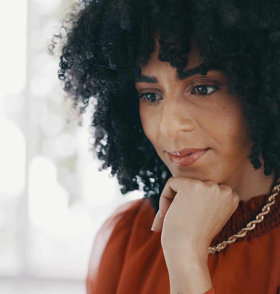Close-up portrait of a woman with curly hair looking down thoughtfully, with her hand under her chin.