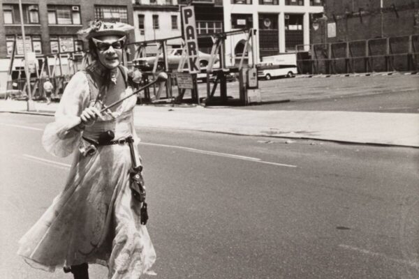 Black-and-white photograph of a person in a flowing dress, hat, and cat-eye sunglasses moving toward the camera on a city street. They hold a thin baton or wand, with buildings, a parking sign, and scaffolding in the background.