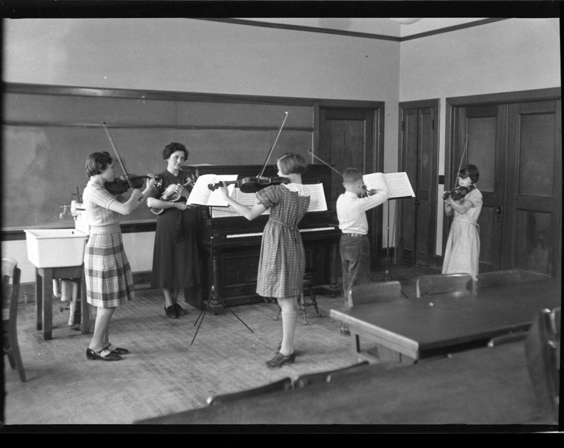 Black-and-white photograph of an instructor standing at the front of a classroom while several students practice violin. A piano, music stands, chalkboard, and classroom tables are visible.