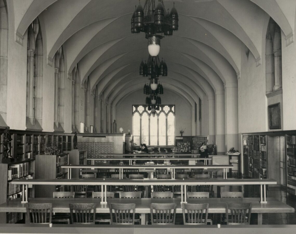Black-and-white photograph of a historic library reading room with long tables and wooden chairs, bookshelves lining both walls, Gothic-style arches, hanging light fixtures, and a large window at the far end.
