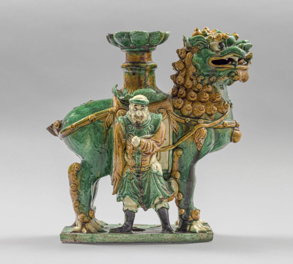 Green and amber glazed ceramic candlestick shaped as a large lion-like creature standing beside a robed human figure. A tall candle holder rises from the animal’s back.