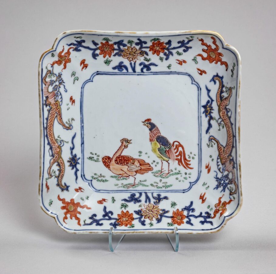 Square porcelain dish with rounded corners, decorated with a colorful rooster and hen at the center. Blue dragons, flowers, and red and green decorative motifs surround the central scene.