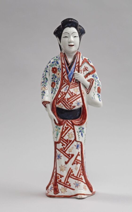 Porcelain figure of a standing woman in a long patterned robe decorated in red, blue, green, and gold. She holds one hand near her chest and wears her dark hair swept upward.