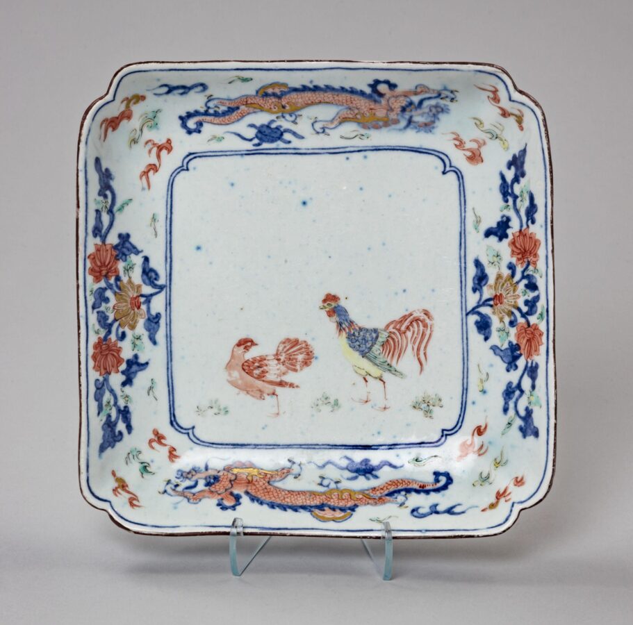 Square porcelain dish with rounded corners featuring a rooster and hen at the center. Blue floral decoration and red dragons fill the border around the birds.