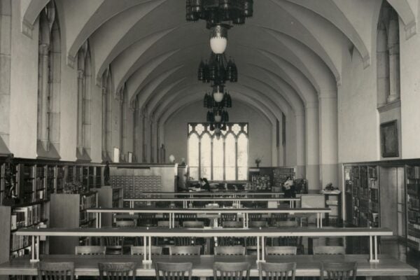 Black-and-white photograph of a historic library reading room with long tables and wooden chairs, bookshelves lining both walls, Gothic-style arches, hanging light fixtures, and a large window at the far end.