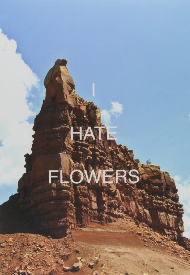 Photograph of a southwest landscape featuring a dramatic reddish rock structure and a blue sky with the words “I HATE FLOWERS” superimposed with white text.