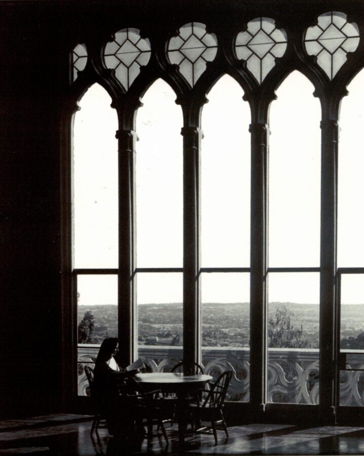 Black-and-white archival photograph of a sister seated at a table, reading beside the same tall Gothic-style window with stained-glass details. The view beyond the window is visible.