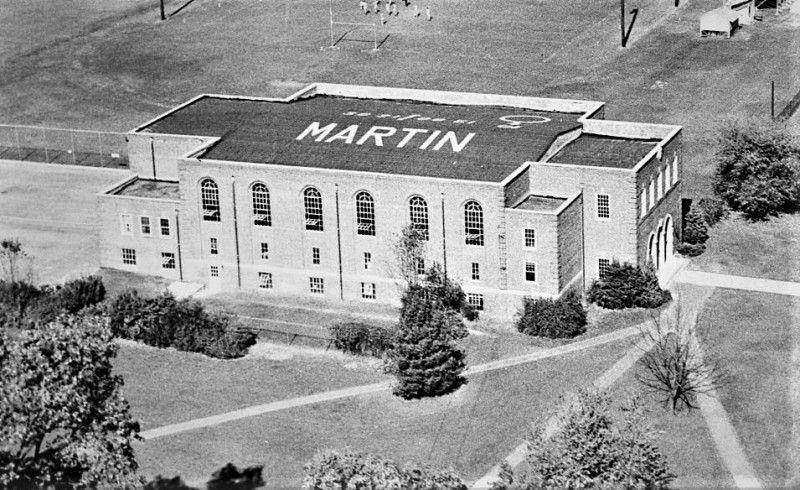 Black-and-white aerial photograph of a large brick building with tall arched windows. “MARTIN” is painted in large letters across its flat roof, surrounded by lawns, trees, and walkways.