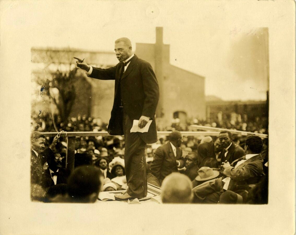 Sepia-toned 1915 photograph of Booker T. Washington, who is dressed in a suit and standing above a large outdoor crowd, gesturing with one hand while holding papers in the other.