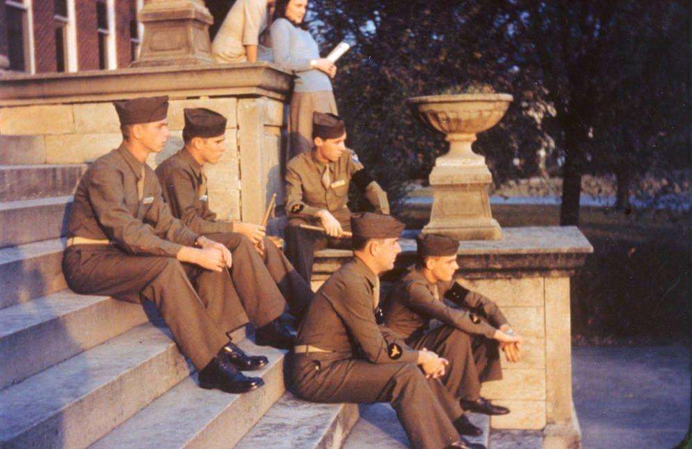 Color photograph of five uniformed cadets sitting on the stone steps of a campus building. A woman stands behind them holding a paper.