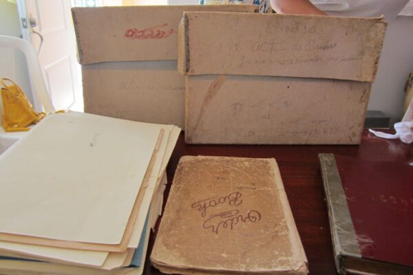 Worn cardboard archival boxes with handwritten Spanish labels sit on a wooden table beside a stack of aged loose papers, a tattered "Order Book" notebook, and a red cloth-bound volume.