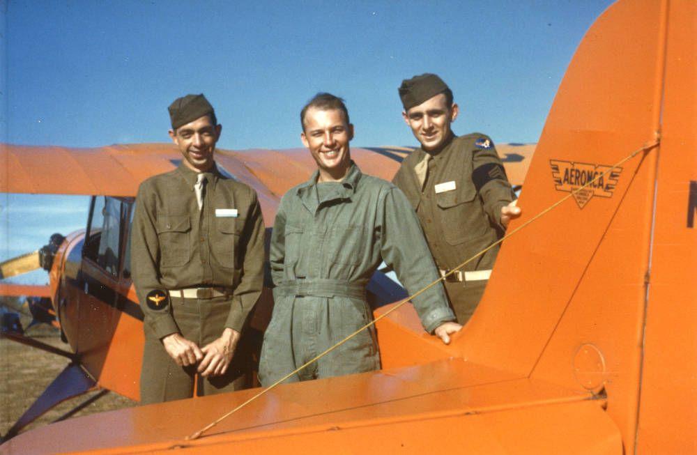 Color photograph of three men standing beside an orange aircraft, with its wing and fuselage filling the foreground.