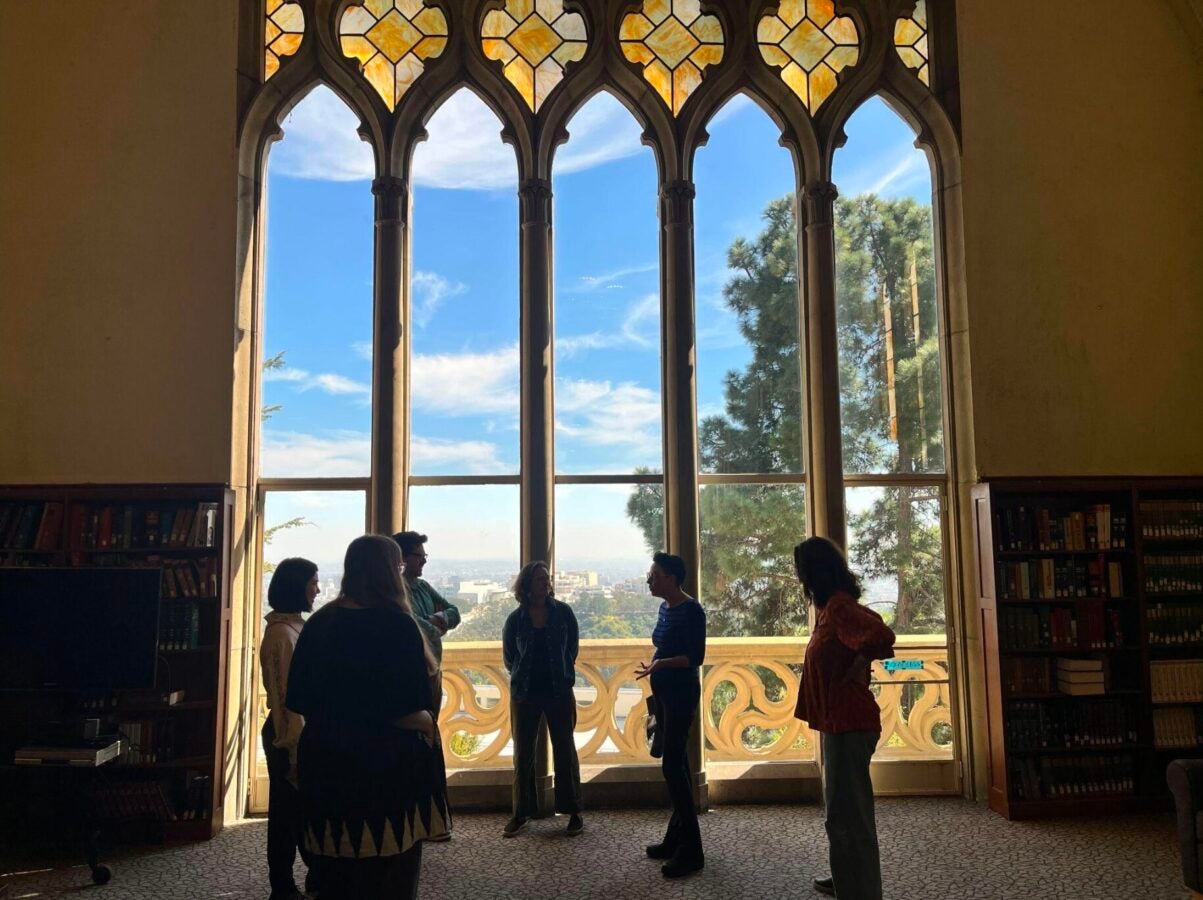 ix people stand and talk in front of a tall Gothic-style window with stained-glass details in a Mount Saint Mary’s University library. Bookshelves flank the window; Los Angeles is visible beyond it.