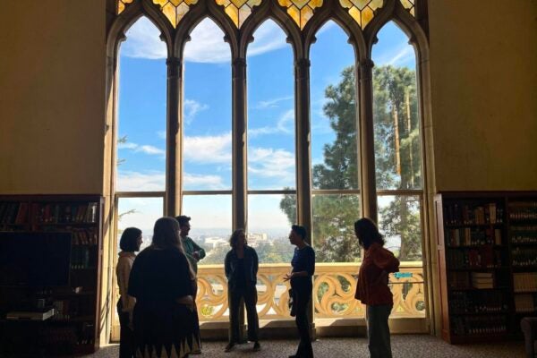 ix people stand and talk in front of a tall Gothic-style window with stained-glass details in a Mount Saint Mary’s University library. Bookshelves flank the window; Los Angeles is visible beyond it.