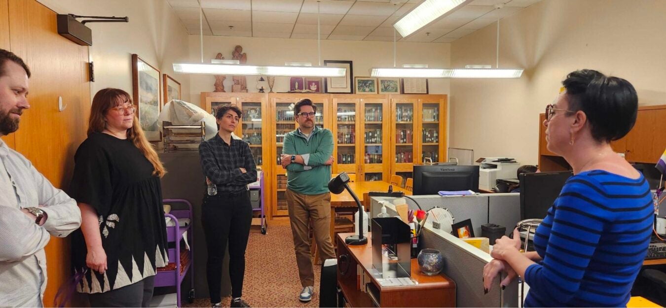 A Mount Saint Mary’s University archivist speaks with four JSTOR and MSMU colleagues in an archives workroom. Shelves, archival materials, equipment, and filing areas surround the group.