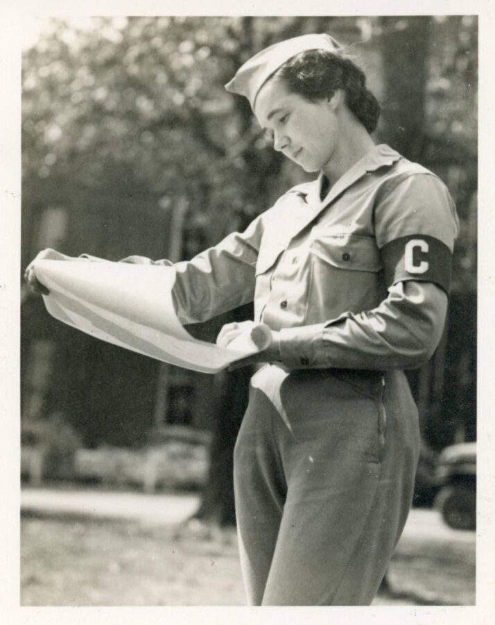 Black-and-white photograph of a woman in uniform and a cap reading a large sheet of paper. A capital “C” appears on her armband.