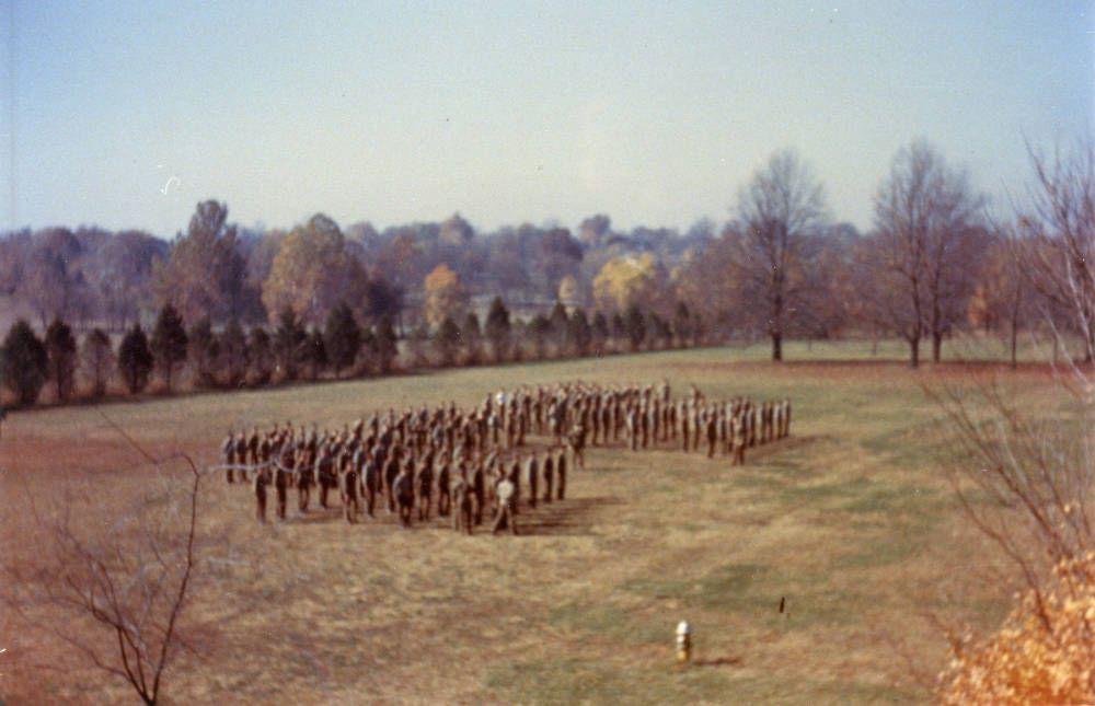 Color photograph of uniformed cadets gathered in formation on a lawn bordered by trees.