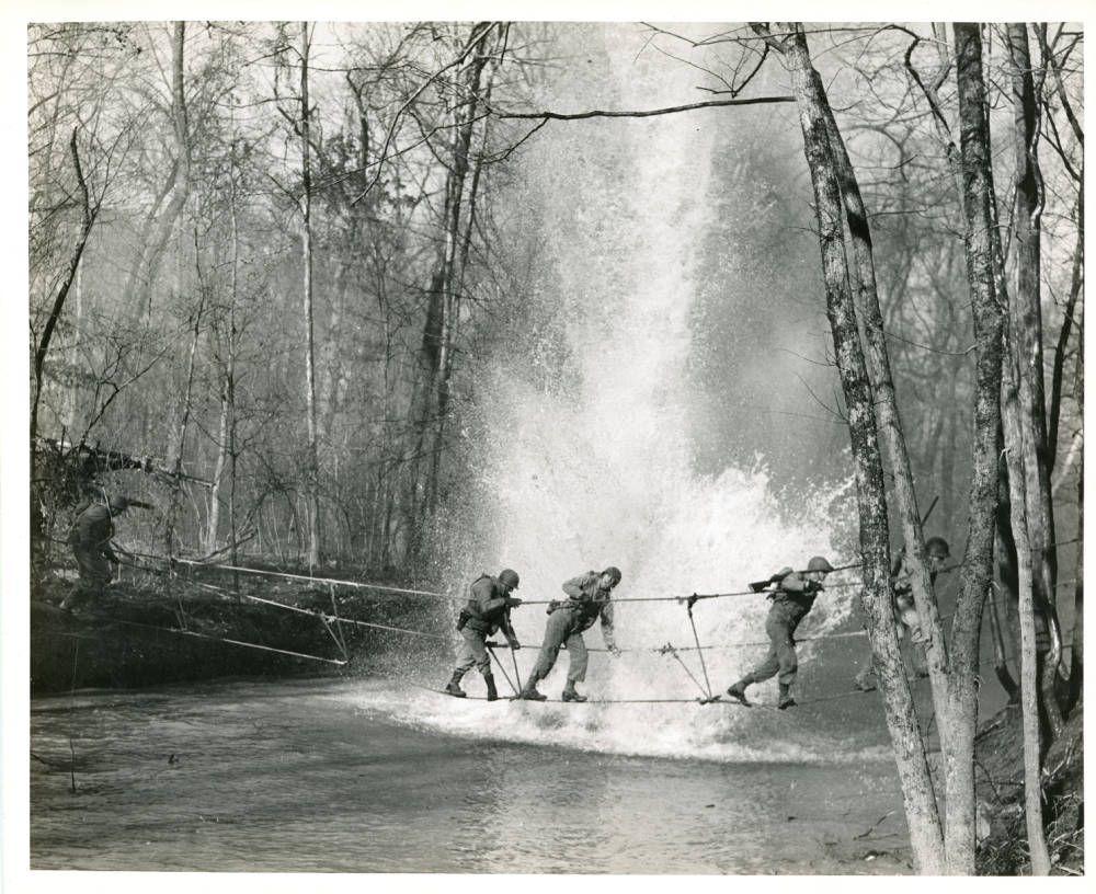 Black-and-white photograph of several helmeted soldiers crossing a rope bridge over a river, with a large splash of water behind them.
