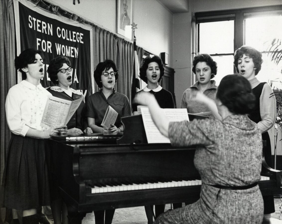 Black-and-white photograph of six women singing beside a piano while a seated conductor leads them, with a “Stern College for Women” banner hanging in the background.