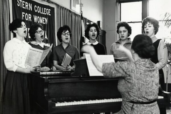 Black-and-white photograph of six women singing beside a piano while a seated conductor leads them, with a “Stern College for Women” banner hanging in the background.