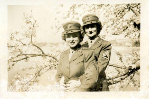 Black-and-white photograph of two women in military uniforms embracing beneath blooming cherry trees.