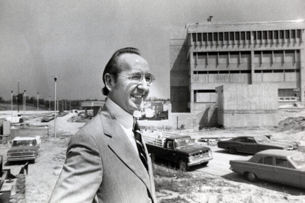 Black-and-white photograph of a smiling man in a suit and glasses standing at a construction site, with vehicles and a large building under construction in the background.