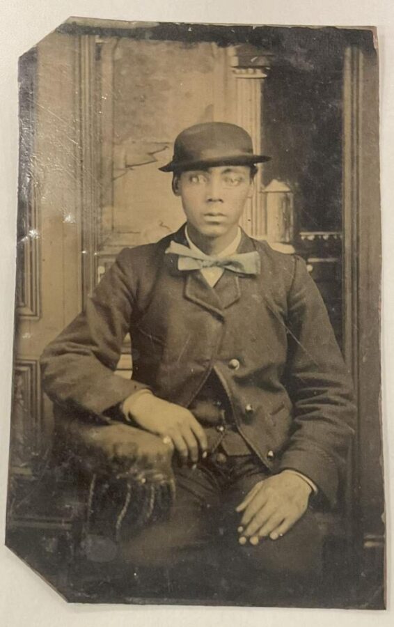 Sepia-toned studio portrait of a seated young man wearing a bowler hat, bow tie, and dark buttoned jacket. He rests one hand on a small table beside him; the aged photograph has visible scratches and worn edges.
