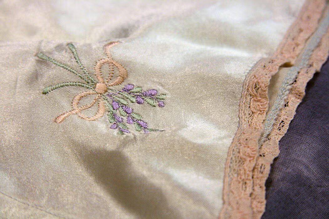 Close-up of pale satin fabric embroidered with a small purple-and-green floral sprig tied with a peach-colored ribbon. Pink lace trim runs along the right edge.