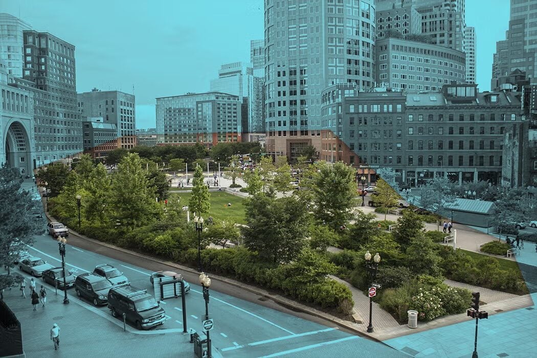 Elevated view of a tree-filled city park bordered by streets with cars and pedestrians, surrounded by dense downtown buildings and skyscrapers.
