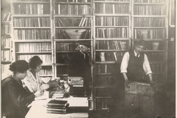 A historical black-and-white photograph of two people working at a table piled with books while two men stand nearby in a library lined with bookcases.