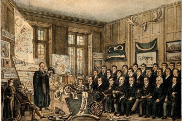 A colored engraving shows a geology lecture at Oxford. A lecturer stands beside a display of maps, rocks, fossils, and a skeleton while rows of male scholars watch from a crowded room. A large mounted antlered skull hangs on the paneled wall above them. The caption reads “The Geological Lecture Room, Oxford.”