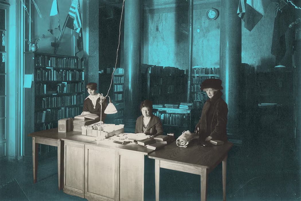 Blue-tinted archival photograph of three women working around a large wooden desk in a library. Two women are seated or standing near books, papers, and filing materials, while a third stands at the right end of the desk. Tall bookshelves line the walls, with columns, hanging flags, and a clock visible in the background.